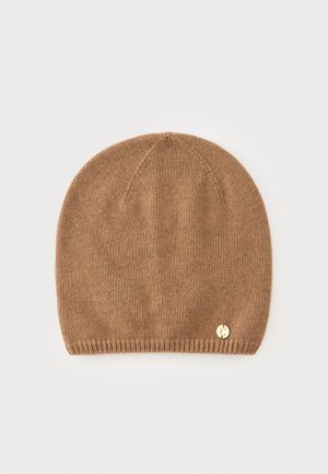 Bonnet en tricot marron avec un petit emblème rond doré près du bord inférieur sur un fond clair uni.