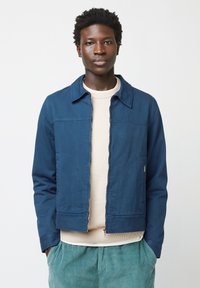 Blauwe ritsjack met kraag, textuurstof, twee zijzakken en een binnenste crême-sweater, gecombineerd met groene corduroy broek.