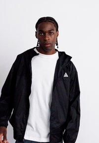 Veste noire légère avec capuche, dotée de poignets élastiques et d'un petit logo blanc sur la poitrine, superposée à un t-shirt blanc et un jean bleu.