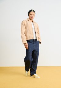 Beige bomber jacket, white t-shirt, and oversized dark denim pants. Yellow sneakers with white accents and black beanie with yellow detail.