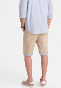 Pantaloni corti beige con orli arrotolati, abbinati a una camicia a righe azzurra chiara. Un orologio visibile e sneaker di colore chiaro completano l'outfit.
