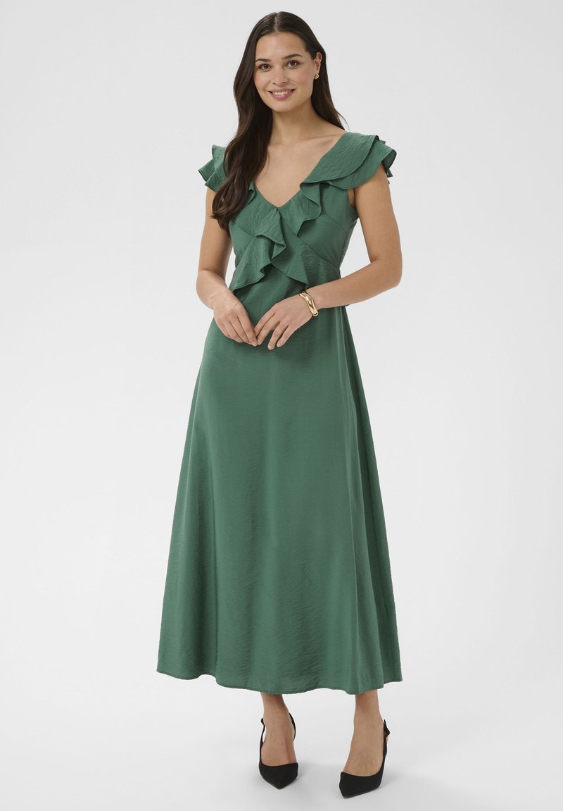 Femme debout portant une robe maxi vert foncé sans manches avec un col en V à volants et des talons noirs pointus, souriant à la caméra.
