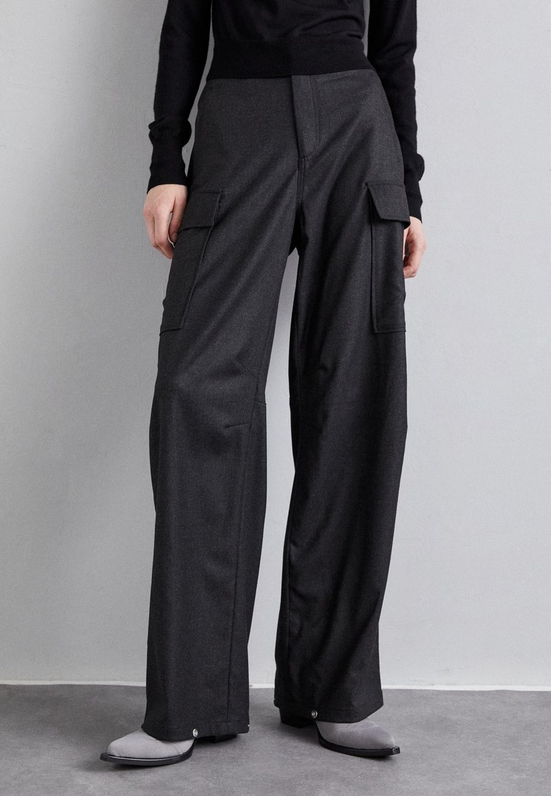 Pantalon cargo noir à jambes larges avec poches à rabat, porté avec des bottes grises et un haut à manches longues noir sur un fond gris uni.