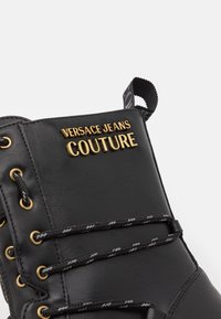 Botte en cuir noir avec œillets dorés, lacets noirs et blancs, et logo doré "Versace Jeans Couture" sur la partie supérieure de la languette.