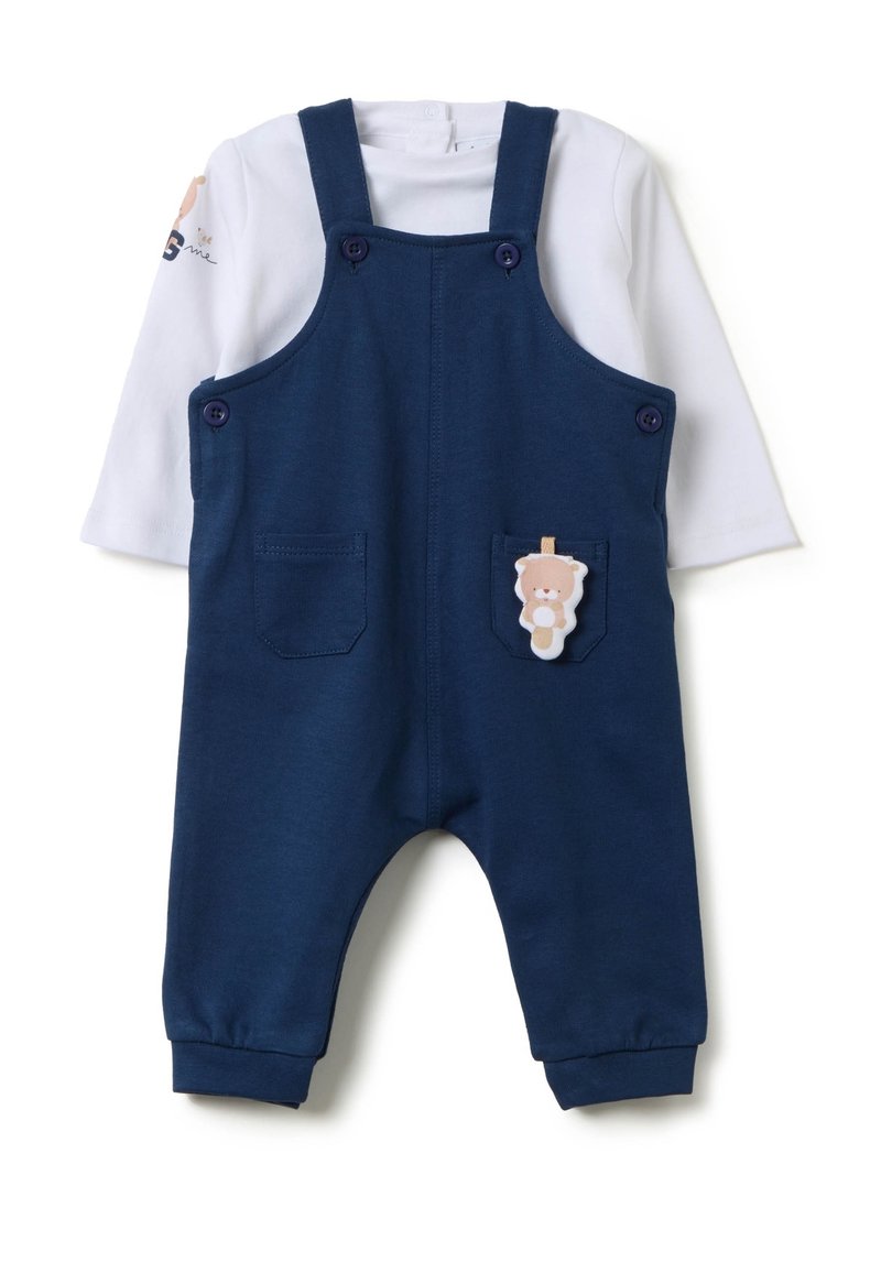 Marineblauer Overall mit verstellbaren Trägern, zwei Vordertaschen und einem Bärenapplikation auf der rechten Tasche. Darunter ein weißes Langarmshirt.