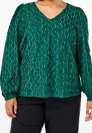 Kobieta w zielonej bluzce z długim rękawem z teksturowanymi pionowymi falistymi liniami i czarnych spodniach, stoi ze zrelaksowanymi ramionami.