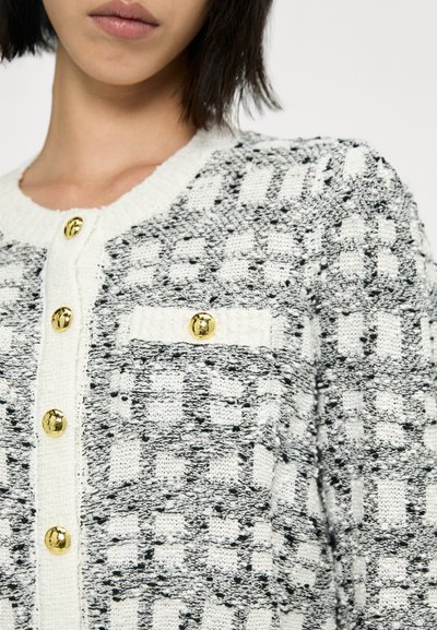 Gilet en tricot noir et blanc avec un motif texturé, doté de boutons en or et d'une petite poche. Vue raccourcie mettant en avant les détails avant.