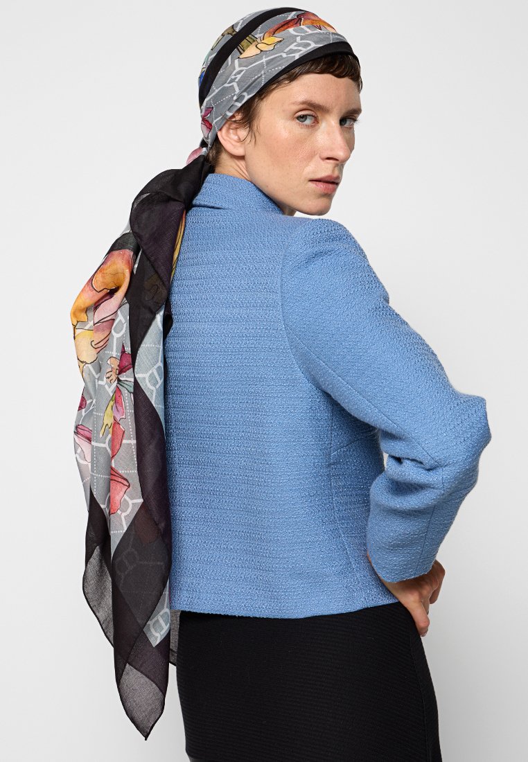 Niebieska kurtka o teksturowanym wykończeniu z dopasowanym fasonem, połączona z kolorowym kwiatowym szalem zarzuconym na ramieniu. Widoczna poniżej czarna sukienka.
