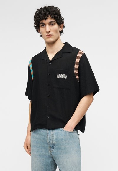 Νεαρός άνδρας με μπούκλες, που φοράει μαύρο κοντομάνικο πουκάμισο με πολύχρωμες ρίγες στους ώμους και το κείμενο «BARROW» στην τσέπη στο στήθος, και μπλε τζιν.