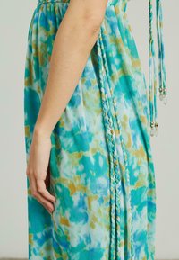 Robe fluide en tie-dye bleu et vert avec un détail tressé sur le côté. Tissu léger, avec des accents jaunes et une finition texturée.