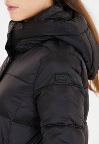Sort pufjakke med høj krave og hætte. Har en quiltet tekstur og et elegant design. Inkluderer et lille mærke på skulderen.