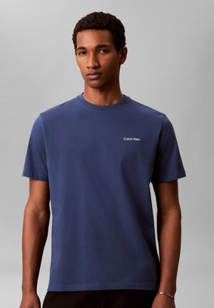 Νέος με κοντά σγουρά μαλλιά που φοράει μια απλή μπλε σκούρα μπλούζα Calvin Klein σε ουδέτερο γκρι φόντο.