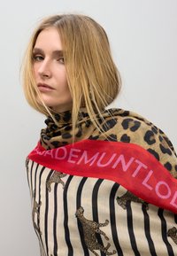 Una bufanda de estampado animal presenta un fondo marrón claro con manchas de leopardo negras y un borde rojo intenso que muestra texto en blanco. La tela parece suave y ligera.