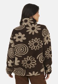 Donna vista di spalle che indossa una giacca in pile marrone con motivi floreali e a spirale beige, abbinata a pantaloni marroni.