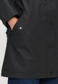 Giacca nera impermeabile con una vestibilità rilassata; presenta una chiusura a bottoni, tasche laterali e una texture liscia.