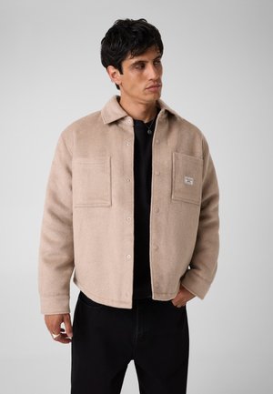 Man met een beige fleecejack met knopen en twee borstzakken, een zwart overhemd eronder en een zwarte broek, kijkt opzij.