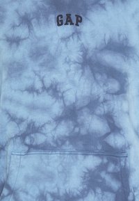 Tissu tie-dye bleu clair avec une poche avant et le logo « GAP » en lettres foncées près du centre supérieur du vêtement.