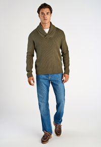 Olijfgroene geribbelde trui met een sjawlboord, gecombineerd met blauwe jeans en bruine veterschoenen. Eenvoudig, textuurvol ontwerp met een casual pasvorm.