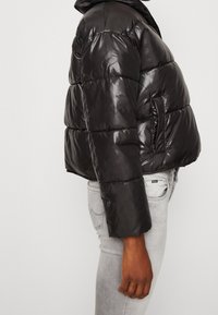 Giubbotto puffa nero realizzato in materiale lucido e trapuntato con cappuccio, dotato di una tasca laterale e di una silhouette aderente. Maniche lunghe.