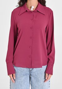 Femme portant une blouse magenta à manches longues avec un grand col et un jean en denim bleu clair.