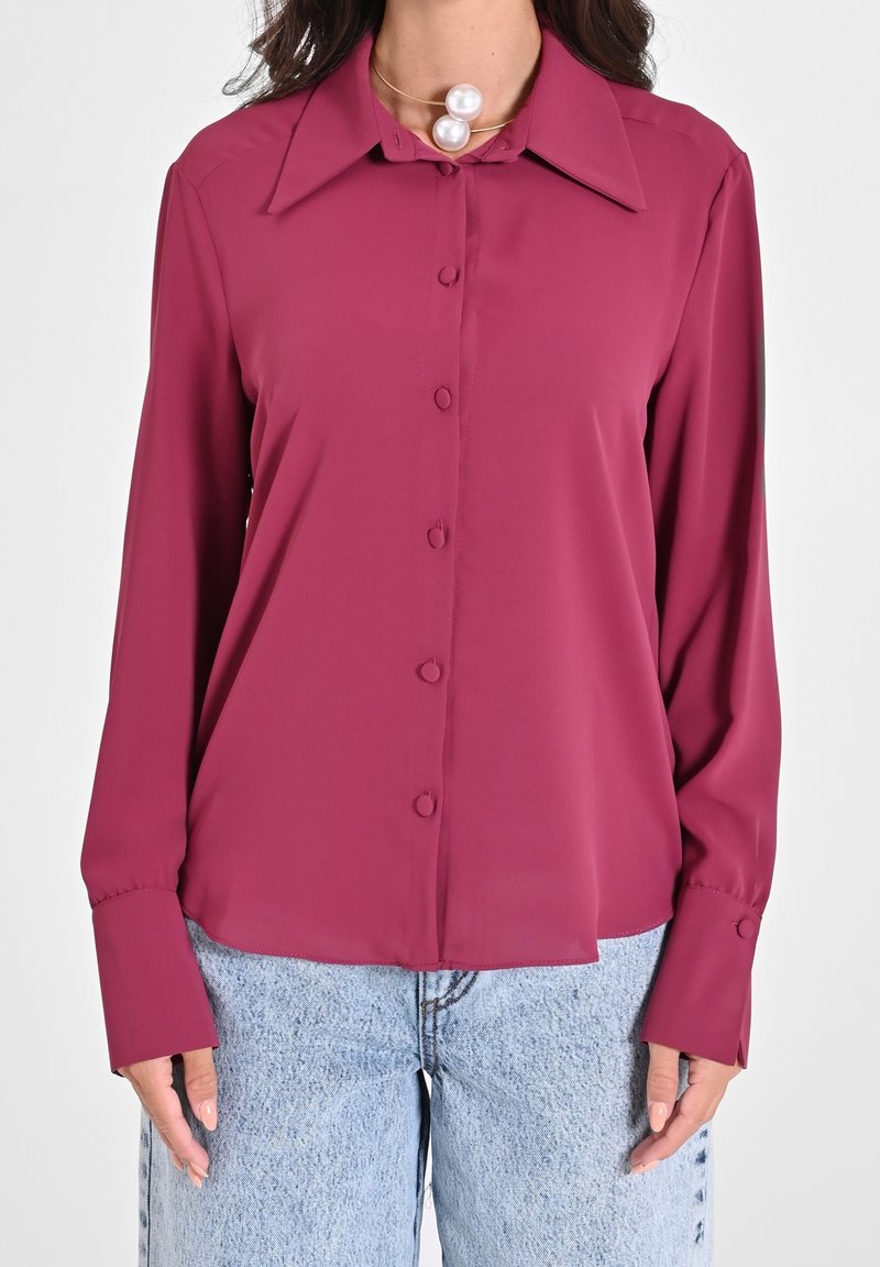 Femme portant une blouse magenta à manches longues avec un grand col et un jean en denim bleu clair.