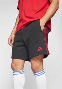 Pantaloni sportivi neri con strisce laterali rosse e logo Adidas rosso, realizzati in tessuto morbido. Indossati con calze bianche con strisce blu.