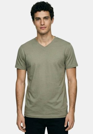 Jeune homme aux cheveux bouclés et foncés portant un T-shirt vert olive à col en V et manches courtes, ainsi qu'un pantalon noir, sur un fond uni.