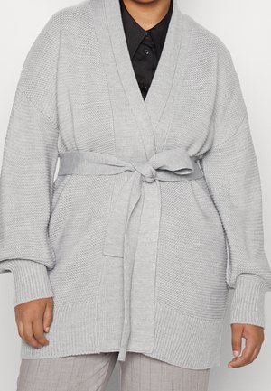 Personne portant un cardigan en maille gris clair avec une ceinture à la taille, sur une chemise noire et un pantalon gris clair.