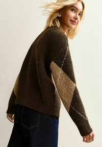 Sweter dzianinowy z brązowym i jasnobeżowym wzorem w romby, o teksturowanym wykończeniu i luźnym kroju. Noszony z ciemnymi dżinsami, z długimi rękawami.