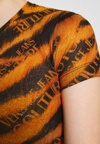 Błyszczący pomarańczowy i czarny materiał z krótkim rękawem, z powtarzającym się napisem "VERSACE JEANS COUTURE", noszony na górnym ramieniu i barku.