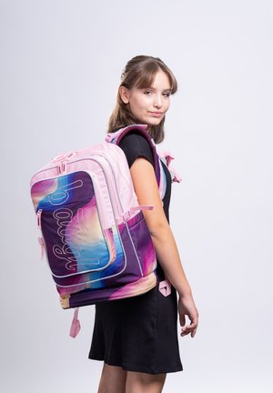 Teenager-Mädchen in schwarzem Kleid mit großem pinkfarbenem und mehrfarbigem Rucksack, seitlich vor einem schlichten hellen Hintergrund posierend.