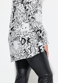 Top bianco con stampe grafiche nere, comprese scritte e volti, abbinato a pantaloni in pelle neri. Il top presenta maniche lunghe e una vestibilità ampia.