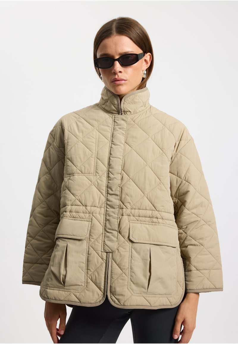 Beige gesteppte Jacke mit hohem Kragen, Frontreißverschluss und zwei großen Taschen. Hat einen lockeren Schnitt und ein strukturiertes Rautenmuster.