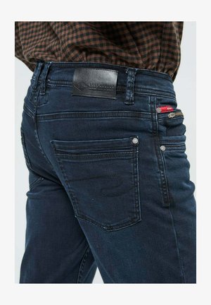 Jean bleu foncé avec poche arrière, poche zippée rouge sur le devant, design cousu sur la poche, porté avec une chemise à carreaux brune.