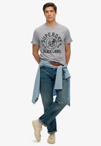 Grijze t-shirt met zwart "Superdry Black Label" graphic, blauwe jeans en een lichtblauwe spijkerblouse die om de taille is geknoopt. Crème kleurige sneakers maken de look af.