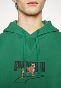 Felpa verde in misto cotone, con un grande logo nero "PUMA" e un design animale ricamato. I lacci sono anch'essi verdi.
