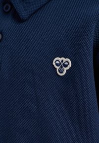 Marinblå bomullspolo med en strukturerad väv. Har en liten ljusbroderad bi-logotyp nära nacken. Två mörka knappar synliga.