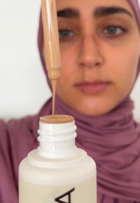 Beige flytande foundation droppas från en pipett ovanför en vit flaska. Bakgrunden är neutral, och pipettens spets är tydligt synlig.