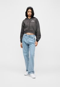 Γυναίκα που στέκεται φορώντας μια κοντή φούτερ New Balance σε σκούρο γκρι, τζιν με ανοιχτό μπλε ρίγες, λευκά αθλητικά παπούτσια και γυαλιά ηλίου στα μαλλιά της.