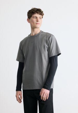 Ung mann iført lagvis grå kortermet T-skjorte over en svart langermet skjorte, stående mot en ensfarget hvit bakgrunn, ser mot høyre.