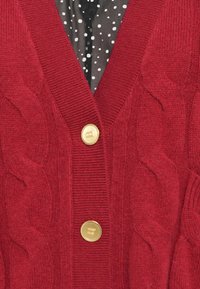 Rød kabelstrikket cardigan med dyp V-hals, tre gullknapper og en svart prikkete bluse synlig under. Teksturert stoff.