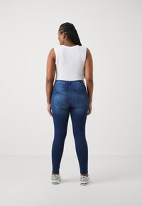 Ταιριαστά σκούρα μπλε τζιν παντελόνια με πίσω τσέπες, συνδυασμένα με ένα κοντό λευκό αμάνικο μπλουζάκι. Το μοντέλο φορά γκρίζα αθλητικά παπούτσια.