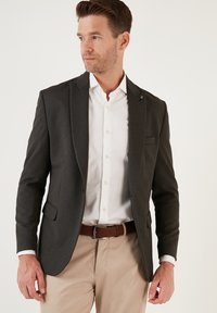 Blazer grigio scuro in tessuto texturizzato, slim fit, con chiusura a un bottone e rever a scapola. Indossato sopra una camicia bianca con colletto.
