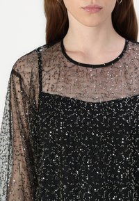 Rochie neagră cu overlay transparent, brodat, care prezintă paiete argintii și modele complicate. Decolteu rotund și mâneci lungi transparente.