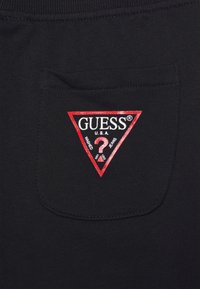 Un bolsillo de tela negra presenta un logo de triángulo rojo con el texto "GUESS" y "U.S.A.", junto con elementos decorativos y un diseño de signo de interrogación.