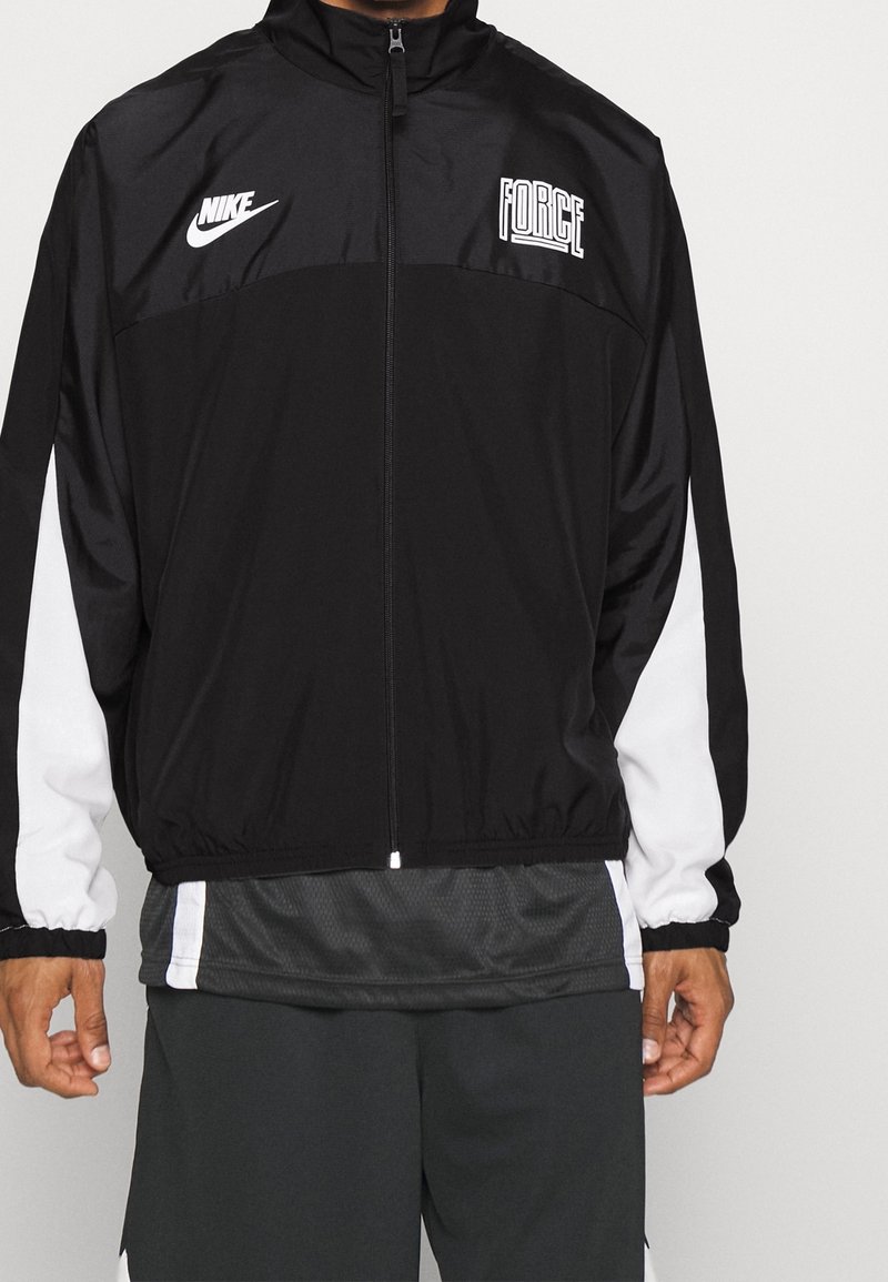 Man wearing black and white Nike zip-up jacket labeled "FORCE," black shorts, standing against a plain light background.