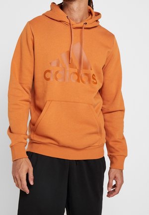 Mann trägt einen orangen Adidas Hoodie mit Kängurutasche und schwarzen Hosen, steht vor einem schlichten hellen Hintergrund.