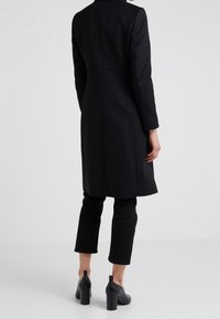 Abrigo de lana negro con corte ajustado, mangas largas y dobladillo recto. Combinado con pantalones cortos negros y botines con tacón.