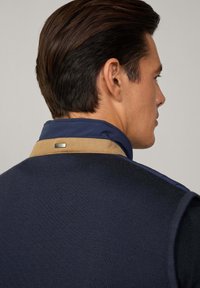 Gilet en tricot bleu marine avec un col beige et bleu marine. Étiquette en métal sur le col. Texture lisse avec des détails côtelés sur le gilet.