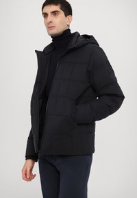Black padded jacket with a hood, featuring a zip closure, side pockets, and a quilted texture. Worn over a black turtleneck.